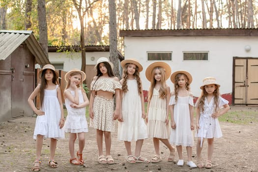 Smiling friendly tween girls in light dresses and straw sun hats standing on courtyard of country house located in pine forest on sunny summer day, enjoying summer vacation