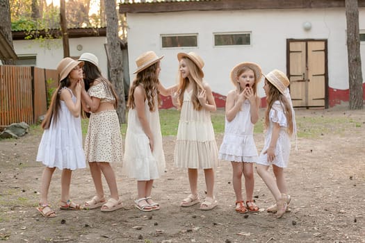 Group of emotional expressive preteen girls in summer dresses and wicker hats chatting on courtyard of country estate. Little gossip girls. Happy childhood and friendship concept