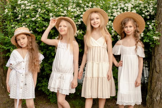 Group of cheerful tween girls in light dresses and wicker sun hats having fun in country estate on summer day, posing in green garden near flowering shrub. Happy childhood