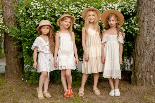 Cute stylish preteen girls wearing light dresses and wicker hats standing near green flowering bush in park on summer day, full-length portrait