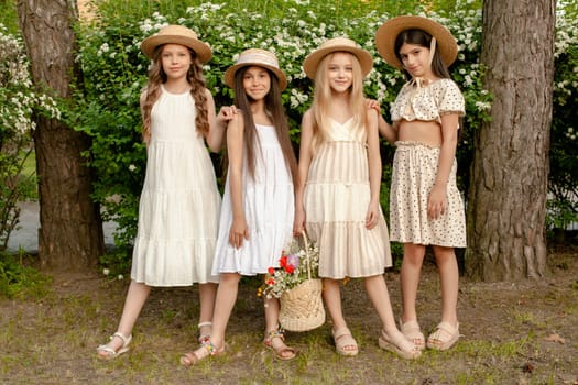 Cheerful friendly tween girls in summer dresses and straw hats holding basket of fresh flowers, posing together near blooming shrub in green park