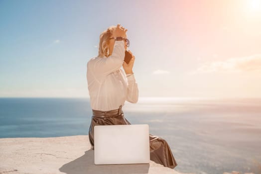 Business woman on nature in white shirt and black skirt. She works with an iPad in the open air with a beautiful view of the sea. The concept of remote work