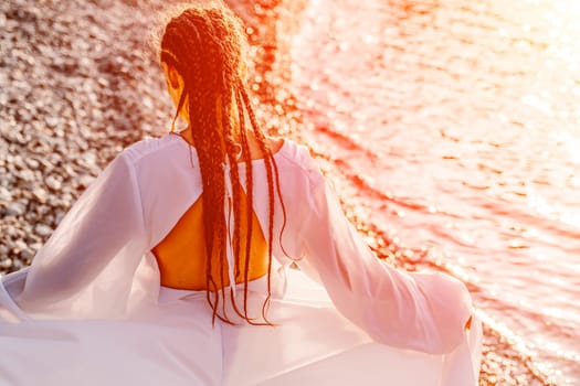 Mysterious woman silhouette long hair walks on the beach ocean water, sea nymph wind listens to the wave. Throws up a long white dress, a divine sunset. Artistic photo from the back without a face.