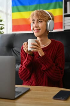 Satisfied young gay man in knitted sweater enjoy morning coffee and listening to music on wireless headphones.