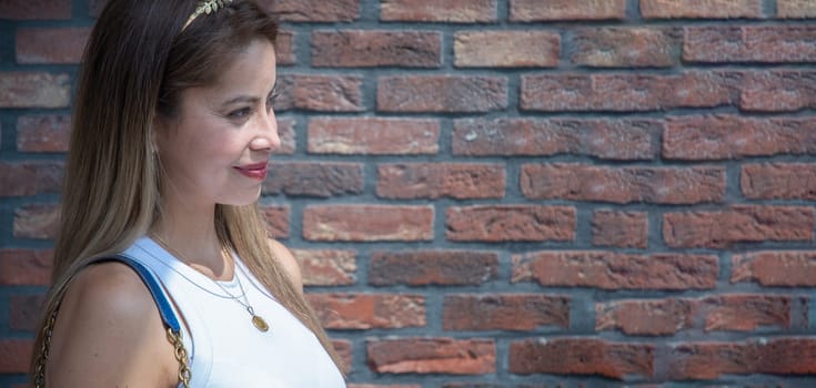 Young blonde woman in stylish clothes looks away smiling near the red brick wall of the house on the street on a bright sunny day, light and shadow, banner with Copy Space, High quality photo