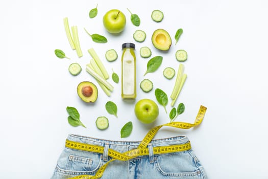 Green fruit, vegetables, smoothie falling into jeans and yellow measuring tape instead of belt on white background. Concept of healthy food for weight loss, detox, diet, healthy clean nutrition