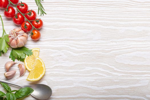 Healthy food ingredients composition with fresh cherry tomatoes, herbs, garlic cloves, lemon wedges and spoon on white wooden rustic background, overhead shot with copy space