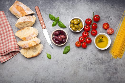 Food composition with sliced ciabatta, olives, olive oil, spaghetti, fresh basil, cherry tomatoes on gray concrete stone rustic background top view, copy space. Italian cuisine concept