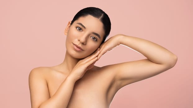 Beauty portrait of multiethnic attractive woman with serene expression holding palms to her face on pink background.