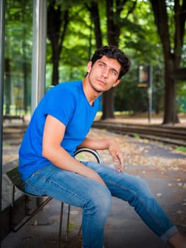 Three quarters shot of one handsome young man in urban setting waiting tram at bus stop, wearing blue polo shirt
