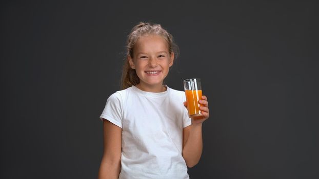 Cute schoolgirl holding a glass of orange juice wearing white t-shirt smiling at camera while standing in studio isolated on black background. Kids fashion concept.