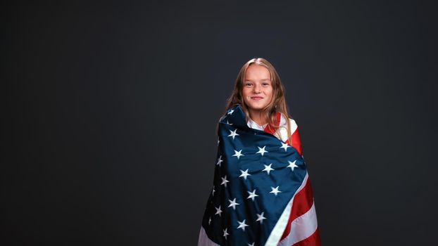Little girl patriot wrapped in a USA flag celebrates independence day expresses patriotism isolated on black background.