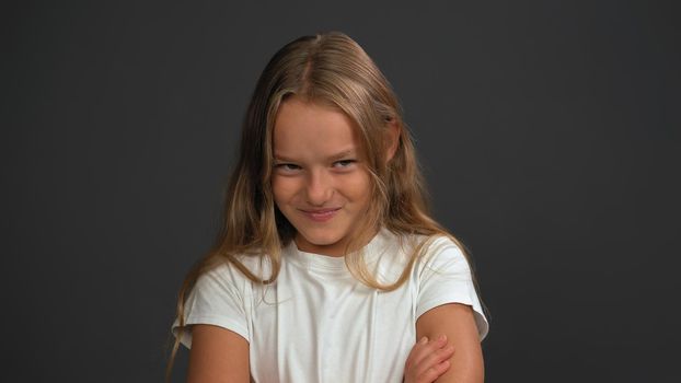 Close up. Little girl of 8,10 years with hands folded looks looks questioningly at the camera wearing white t shirt isolated on dark grey or black background.