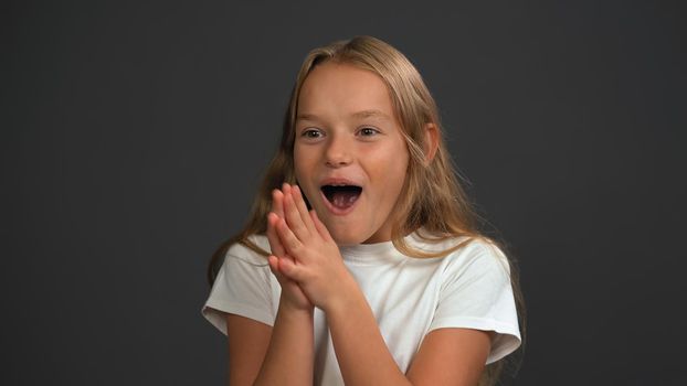 Excited little girl standing with hands put together happily looking a side of camera, wearing white t-shirt isolated on dark grey or black background.