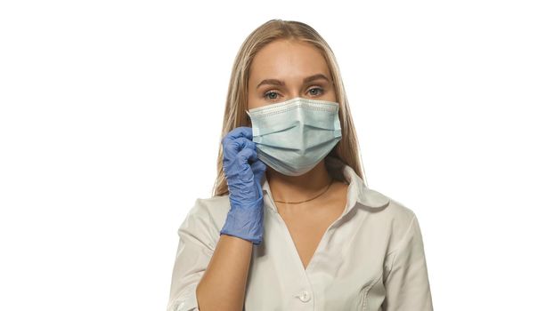 Putting on a face medical mask young blond nurse looking at the camera, wearing white medical uniform and disposable blue rubber glows isolated on white background.