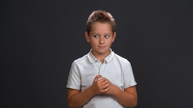 Frustrated boy in white shirt holding hands together in front of him while looking sideways at upper corner. Isolated on black background.