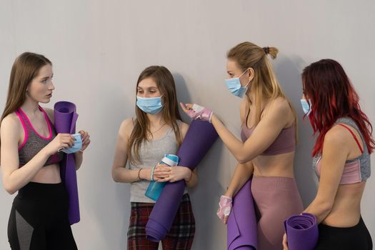 Convincing friend to wear medical face protective mask athletic girls in sports out fits standing next to the wall holding a yoga mat, talking to each other, standing at a grey background.