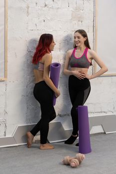 Beautiful young women in sports out fits talking standing next to the wall holding a yoga mat, smiling to each other, standing on a white brick wall background.