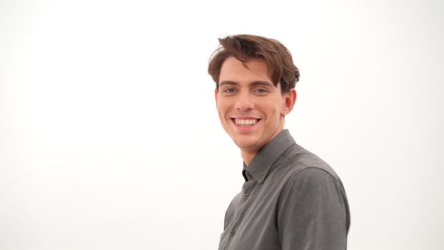 Smiling handsome young man turn fixing his hair and looking at camera wearing grey shirt isolated on white background. 4K video.