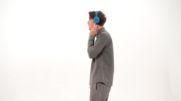 Young guy in grey duffle or outfit dancing standing sideways listening music wearing blue wireless headphones. Funny dancing young guy listen to his favourite track or song. 4K footage.