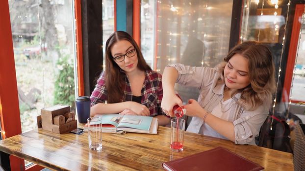 Drafting drink in glasses girl friends enjoying fresh made juice together in a cafe with lights running on the background as they sit at a table chatting and laughing. 4K footage.