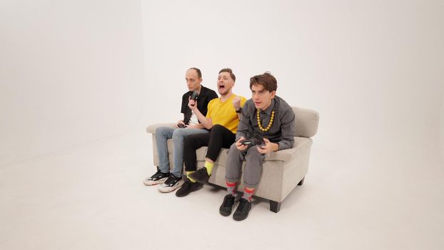 Happy winning while gaming guy in yellow shirt playing video games with friends sitting on a white sofa isolated on white background. Guy reacts to winning or a lose. Gaming concept. 4K footage.