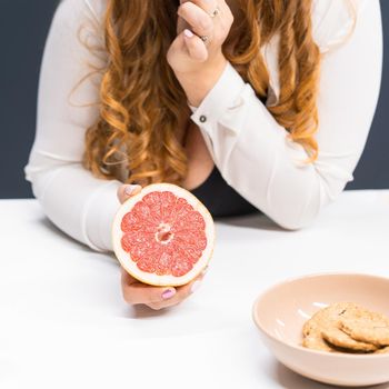 Fat young woman holding a fresh grapefruit in hand standing leaned on a kitchen table at home with long curly blond hair. Dieting and nutrition concept. Square cropped.