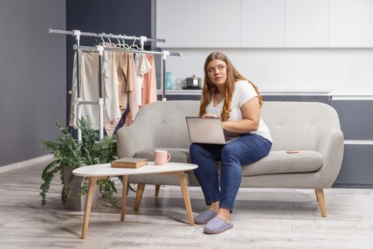 Wondering looking at the window young fat girl working on laptop sitting on the sofa at home with the kitchen on background. Working distantly from home using laptop.