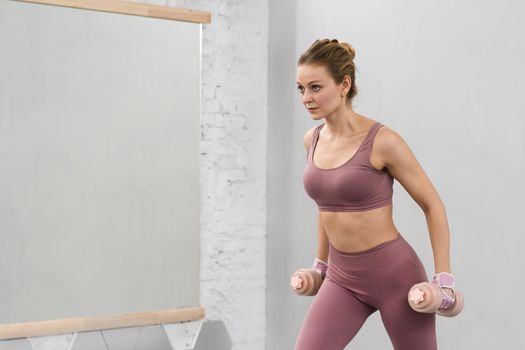 Girl in tight pink outfit working out with dumbbells. Beautiful fitness girl training legs and lifting weights. Girl doing sports in a loft interior studio. Healthy lifestyle concept.