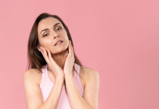 Moisturising her face beautiful woman with no makeup standing with hands on her chicks, massaging it gently attractive brunette girl on pink background. Skin care concept.