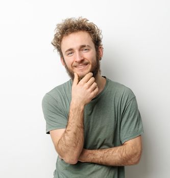 Curly hair smiling handsome young man looking at camera touching chin dressed in olive t-shirt isolated on white background. Portrait of smiling young man with hands folded.
