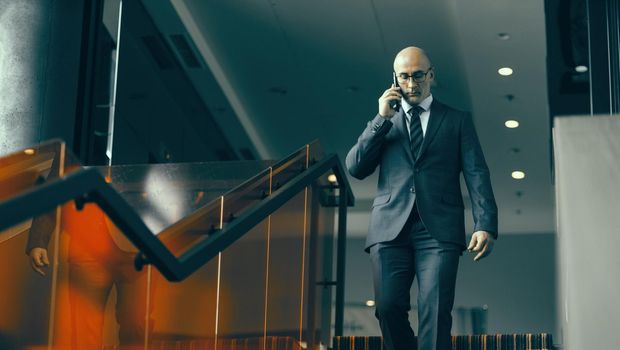 Walking down the stairs and having a conversation on the phone mature bald businessman. Handsome concerned man walking down modern office stairs.