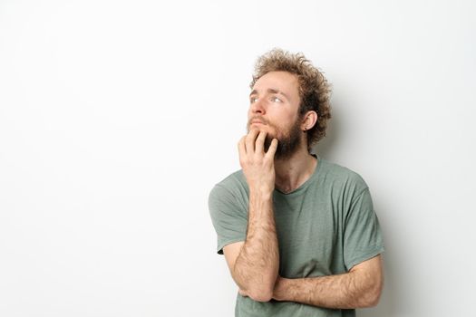 Pensive scratching beard young man with hands folded. Handsome young man with curly hair in olive t-shirt looking up away isolated on white background.
