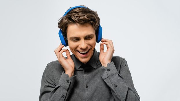 Joyful guy in grey shirt listening music wearing blue wireless headphones enjoying sound with closed eyes. Happy young guy listen to his favourite track or song.