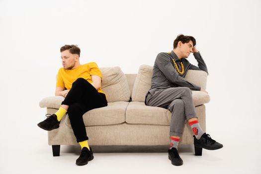 Quarreled guys are sitting on opposite sides of the sofa. Two trendy young mens are sitting on a soft couch and not communicate isolated on white background.