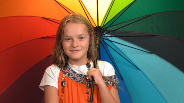 Charming little girl in orange dress holds rainbow colors umbrella standing and cheering in studio while looking at the camera.