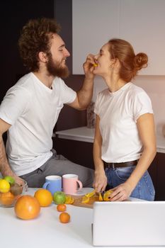 Feeding orange to girl young man embracing each other enjoying their their morning coffee in a new house. Making fun at modern kitchen and kissing while cooking at home. New house concept.