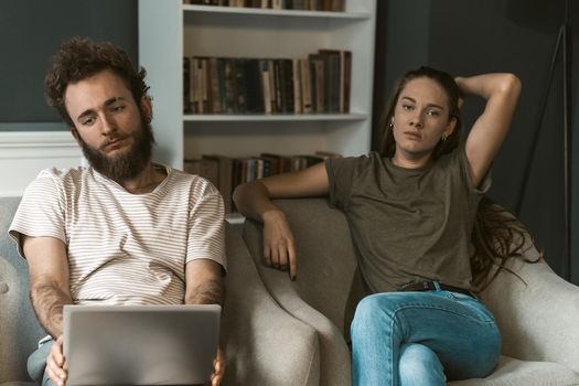 Young couple had a fight staying quarantined at home with sitting quiet on the arm chairs. Not happy young couple sitting on a chair together using laptop.