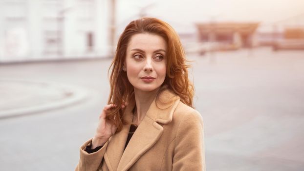 Beautiful woman standing outdoors posing for camera in an autumn beige coat, beautiful hair with urban city background. Vertical portrait. Warm tinted photo.