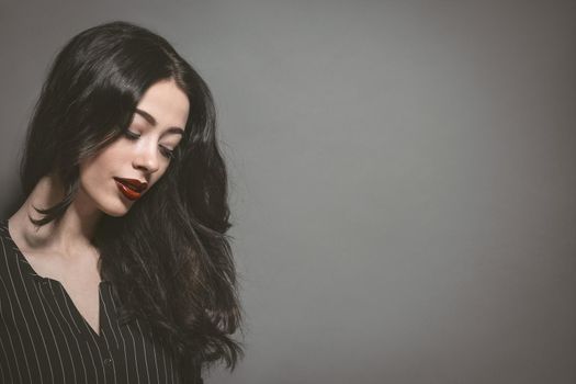 Portrait of young woman with long brunette hair and big lips in a black striped jacket isolated on gray background. Young female model with professional make-up. Cosmetics. Fashion, beauty concept.