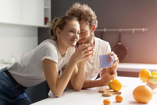 Morning coffee in a new home gently kissing woman young man making breakfast in a new modern kitchen and smiling to each other. Pretty girl feeds or nursing her boyfriend.