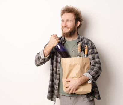 Back from shopping holding a paper bag with bottles of wine smiling young man. Handsome young man with curly hair in olive t-shirt looking at camera isolated on white background.