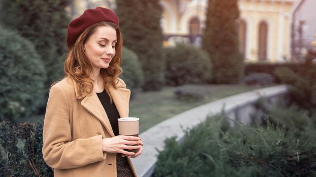 Beautiful young woman holding a mug with coffee standing outdoors. Portrait of stylish young woman wearing autumn coat and red beret outdoors. Autumn accessories.