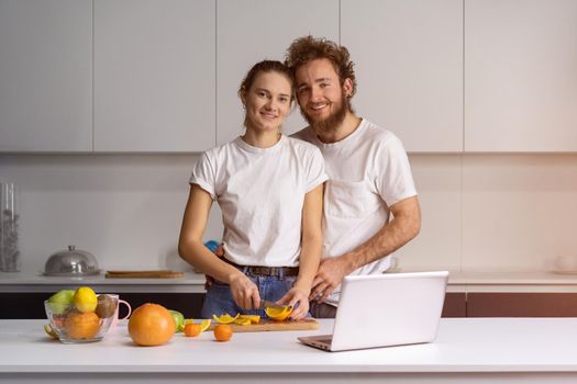 Happy family in their new house. Handsome man hugging wife smiling looking at camera. Beautiful young couple talking on video call using laptop. Young couple cooking healthy food in kitchen at home.