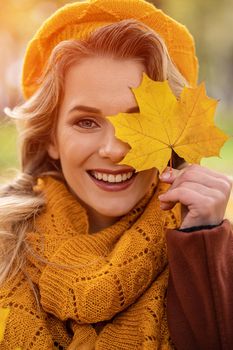 Happy young girl cover her eye with a yellowed leaves in yellow knitted beret with autumn leaves in hand and fall yellow garden or park. Beautiful smiling young woman in autumn foliage.