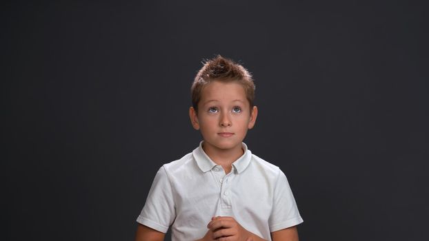 Expecting miracle boy in white shirt holding hands together in front of him while looking sideways at upper corner. Isolated on black background.