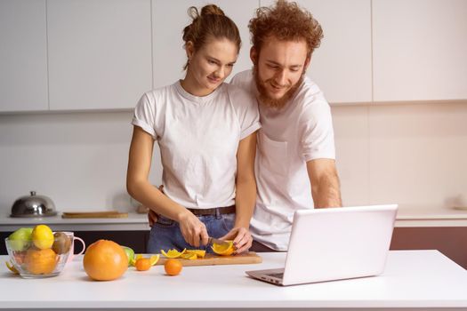 Man leaned on girl smiling watching romantic movie. Beautiful young couple talking on video call using laptop. Young couple cooking healthy food in kitchen at home.