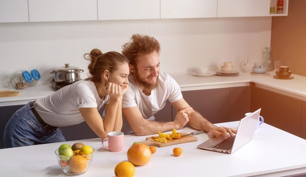 Young couple cooking healthy food in kitchen at home. Girl leaned on man smiling watching romantic movie. Beautiful young couple talking on video call using laptop.