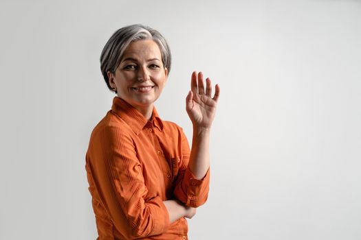 Happy middle aged grey haired woman smile looking at camera with one hand lifted up in a folded arms wearing orange shirt isolated on white background. Human emotions concept.