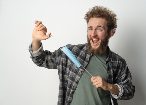 Fooling around playing with protective medical face mask young handsome man holding it stretching it in front of face wearing plaid shirt and olive t-shirt under. White background.